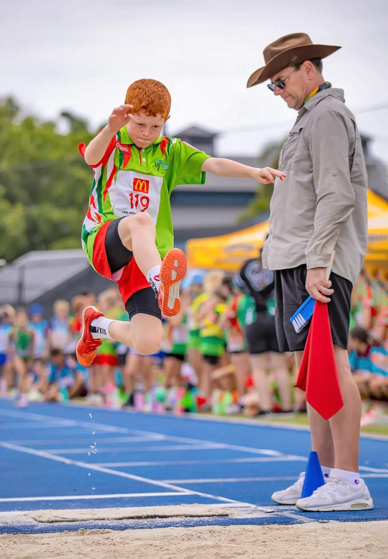 Young boy mid-air during a long jump at a track and field event, with an official watching nearby