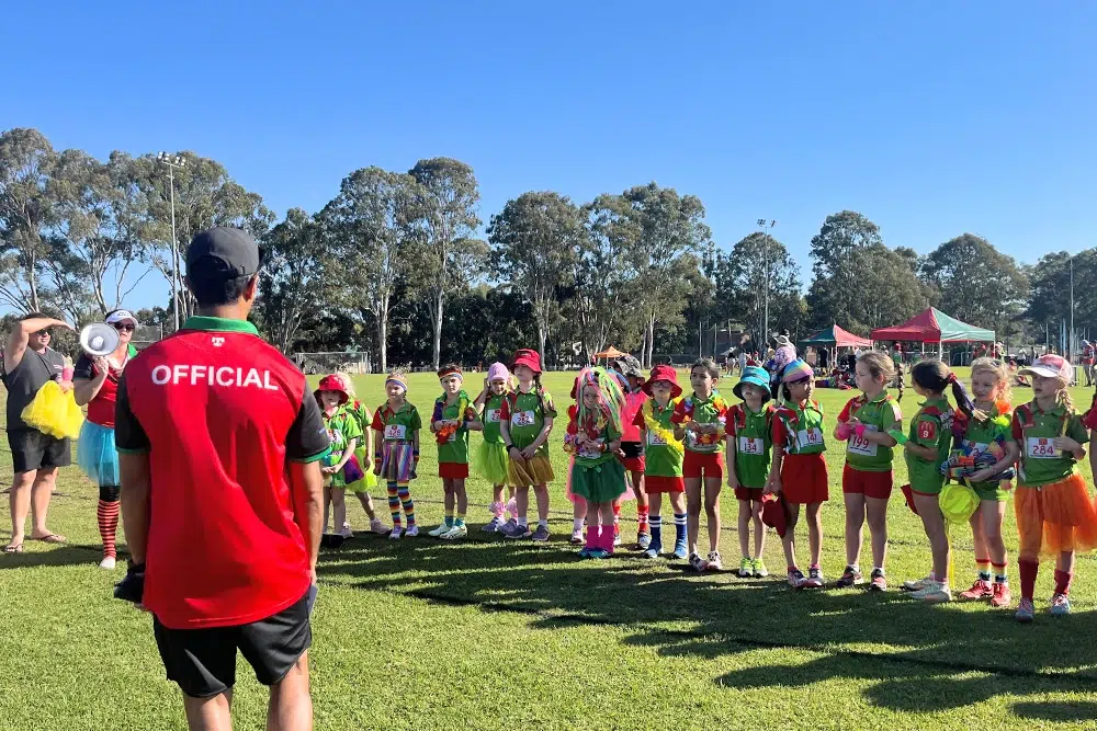 A group of children in colorful costumes stand in a line on a grassy field, facing an event official wearing a red "OFFICIAL" shirt, during a sports or fun day event under a clear blue sky.