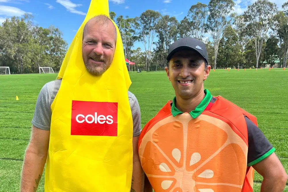 Two men standing on a grassy sports field, one wearing a yellow banana costume with a "Coles" logo, and the other dressed as an orange slice, smiling at the camera.