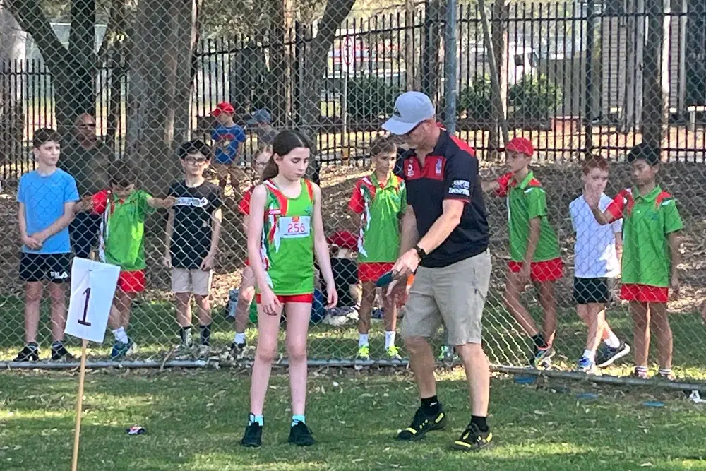A young girl in a green and red athletic uniform stands near a coach who is giving instructions, with other children watching behind a chain-link fence.