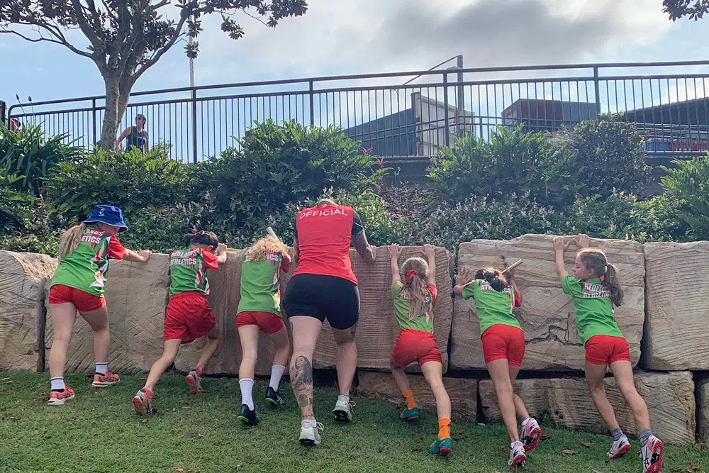A group of young girls in green and red "Redlands Athletics" uniforms and an official in a red shirt lean against a large stone retaining wall, appearing to stretch or climb during a track and field event.
